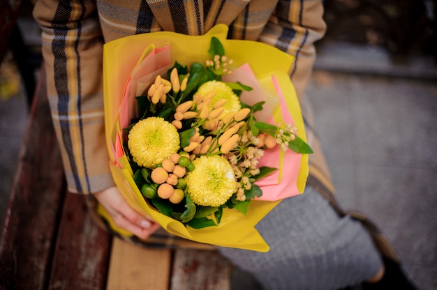 Top view of woman in plaid coat sitting on bench with a bouquet of yellow flowers