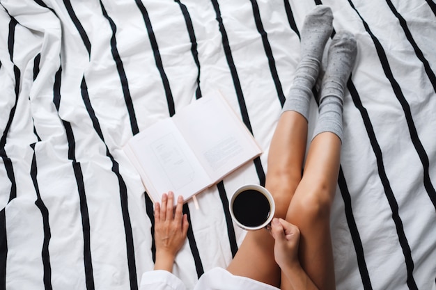 Photo top view  of a woman reading book and drinking hot coffee in a white cozy bed at home in the morning