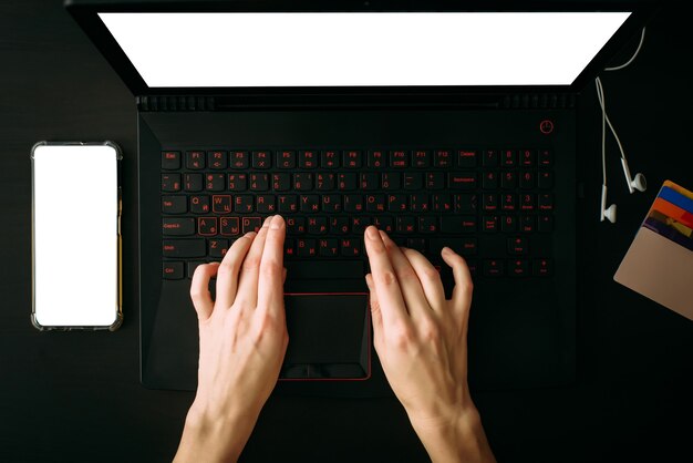 Top view woman's hands typing on laptop  with white blank screen. Empty screen smartphone, headphones, credit cards on black table