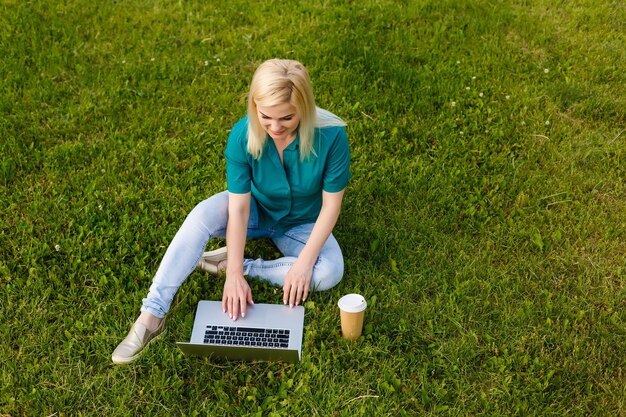 ラップトップ、キーボードの手で緑の芝生の公園に座っている女性の上面図。コンピューター画面のモックアップ。屋外で勉強している学生。テキスト用のスペースをコピーする