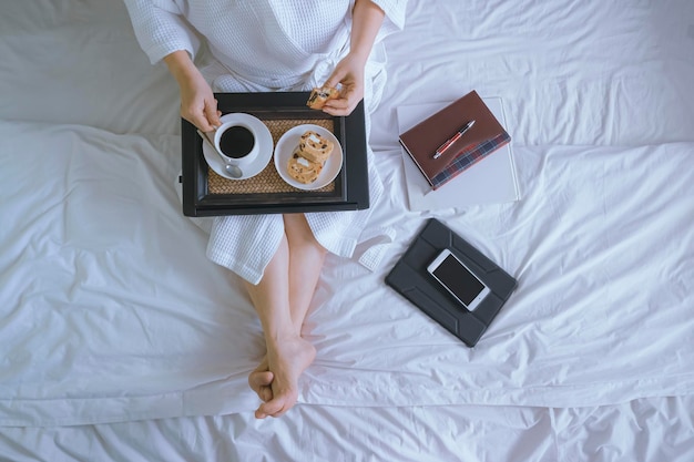 Top view of woman in white bathrobe drinking coffee with breakfast after work online on the bed