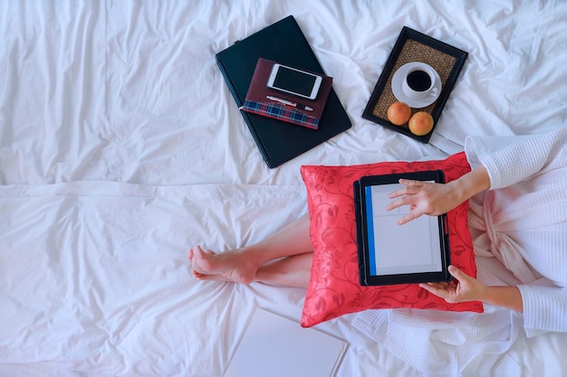 Top view of woman in white bathrobe using digital tablet with morning coffee on her bed