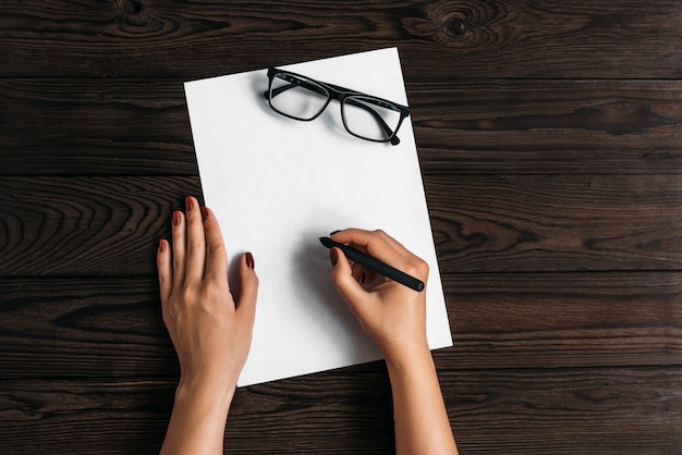 Top view of women's hands, ready to write something on an empty piece of paper lying on a wooden table.