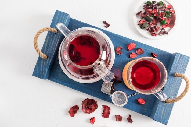 Top view of a wooden blue tray with a glass teapot and a cup of\
red hibiscus tea healthy tea for breakfast