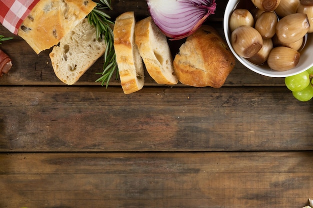 Top view of a wooden cutting board with bowls of snacks, some garlic, a bunch of grapes and slices of bread, arranged on a on a textured wooden surface.