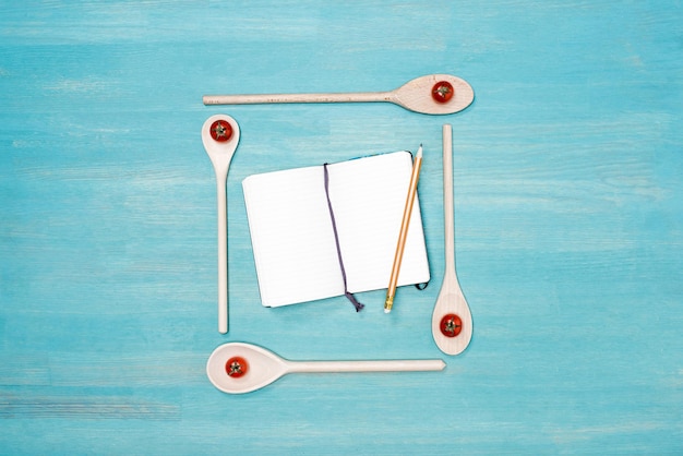 Top view of wooden spoons with cherry tomatoes and blank open cookbook with pencil on table