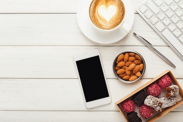 건강한 생채식 사탕, 커피 컵, 아몬드 그릇, 스마트폰, 노트북 키보드가 있는 작업 테이블의 최고 전망. 사무실에서 피트니스 디저트, 복사 공간