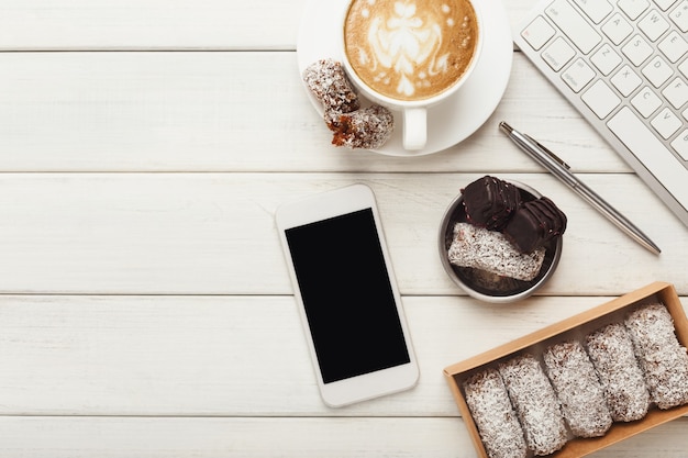 Top view on working table with healthy raw vegan candies, coffee cup, smartphone and laptop keyboard. Fitness dessert at office, copy space