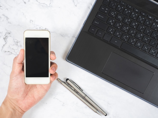 Top view workspace hand holding mobile phone with silver pen and laptop white marble desk