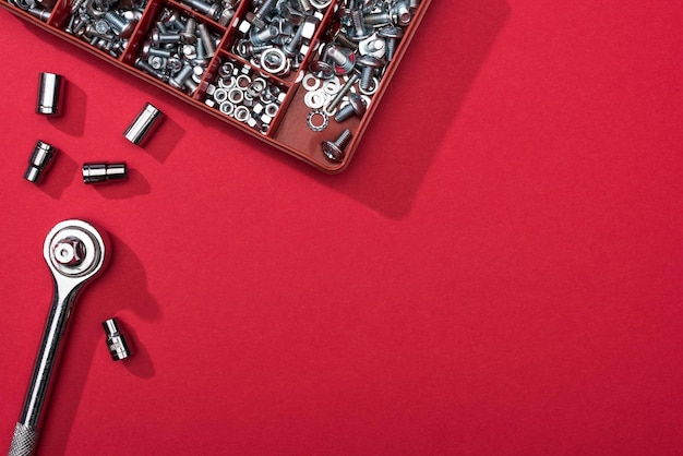 Вид сверху на гаечный ключ с насадками и ящик для инструментов с болтами и гайками на красном фоне