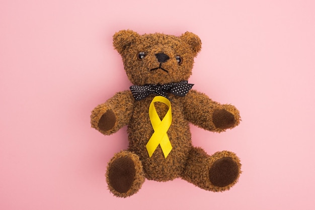 Top view of yellow awareness ribbon on teddy bear with bow on pink background international