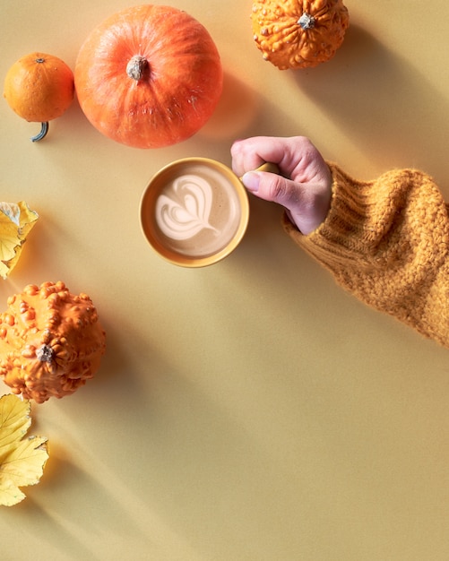 Top view on yellow Fall still life with latte in ceramic mug on yellow paper. Yellow paper flat lay with hand with cup of coffee, orange pumpkins, quince and pair of canvas sneakers.