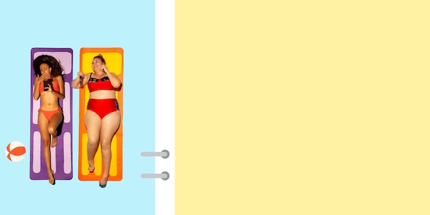 明るい水着の海と砂のように青と黄色の背景にビーチリゾートで休んでいる若い白人モデルの上面図。コピースペース。夏、パーティー、寒さ、休暇、友情の概念