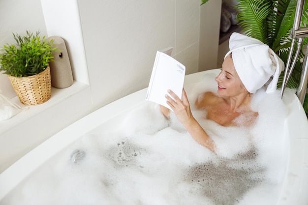 Vista dall'alto sulla giovane bella donna caucasica che si rilassa nella vasca da bagno con schiuma e libro di lettura