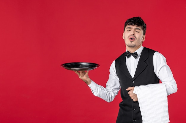 Top view of young dissatisfied man waiter in a uniform with bow tie holding tray and towel