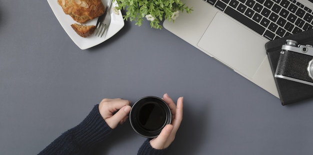 Top view of young female holding coffee cup in winter workspace with office supplies on grey desk 