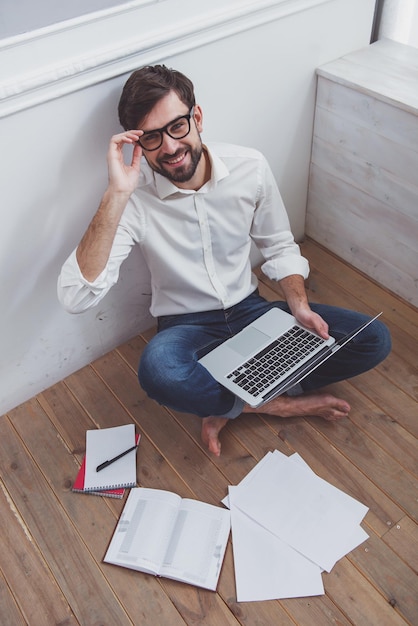 Top view of a young handsome businessman in white classical shirt jeans and eyeglasses using a laptop smiling sitting on the wooden floor and working