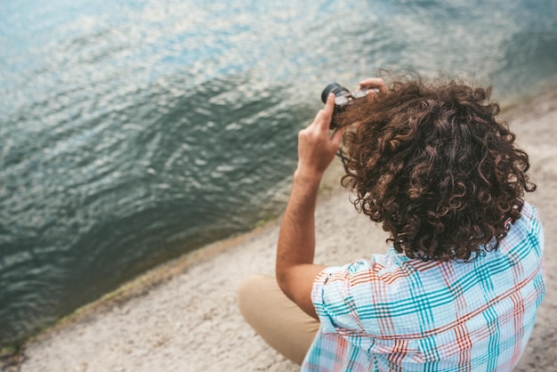 彼のデジタルカメラで自然の写真をチェックする巻き毛を持つ若い男性の上面図若いハンサムな男は、湖の横でポーズをとってデジタルカメラでカジュアルなシャツを着ています人々はライフスタイルを旅行します