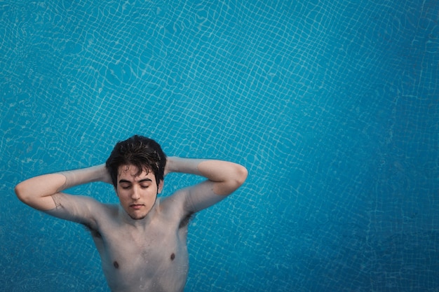 彼の頭の後ろに彼の手でプールに浮かんでいる若い男の上面図。ホテルで日光浴をしながら休暇を楽しんでください。休日の概念。