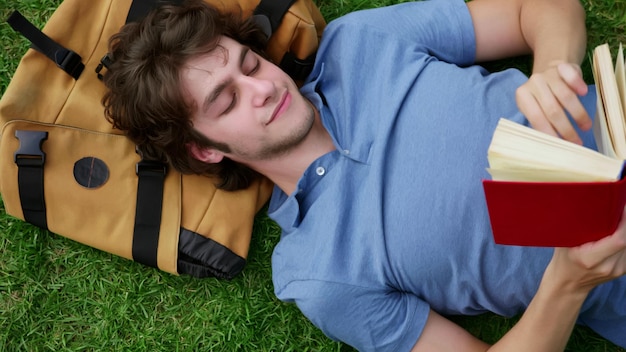 Top view of young man lying on grass reading book with head on backpack