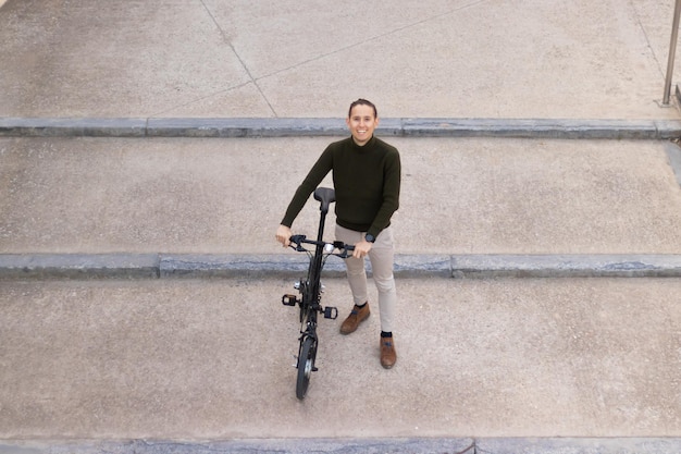 Top view of young man smiling to the camera going down the\
concrete steps with a bicycle on his hands city life concept