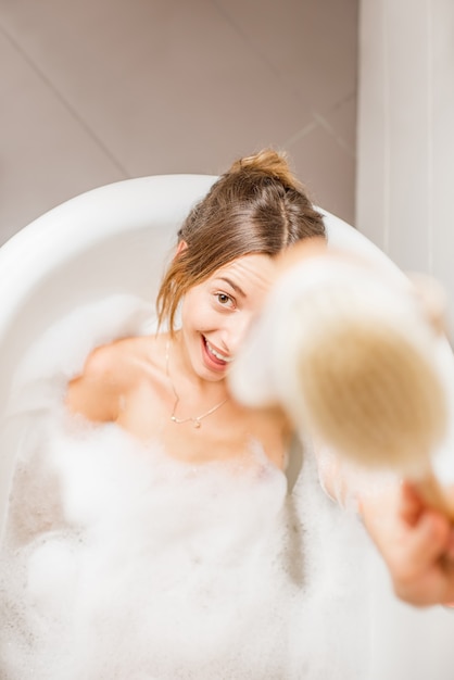 Top view on the young playful woman lying with brush in the bath full of foam