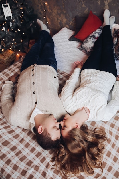 Top view of young pretty woman and handsome man in white sweaters lying on bed while holding hands.