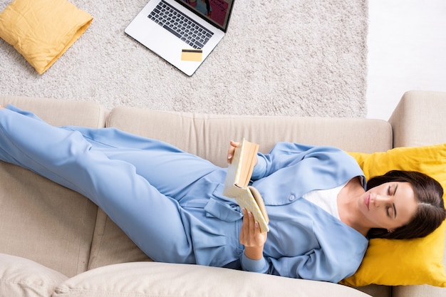 Top view of young restful woman in blue pajamas holding open book in front of herself while lying on soft comfortable couch in living-room