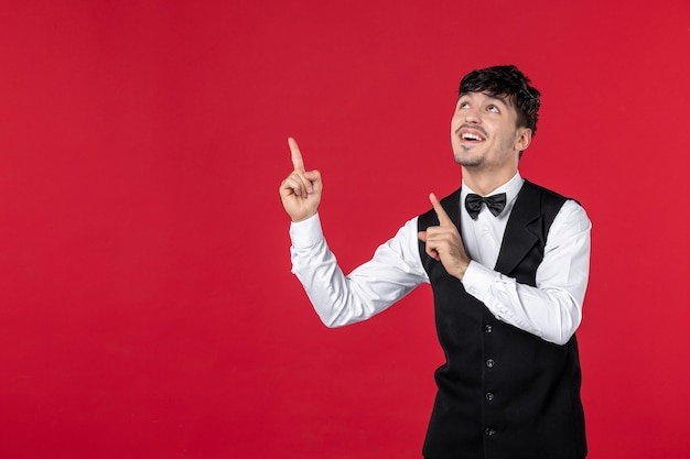 Top view of young smiling male waiter in a uniform with bow tie and pointing up on the right side