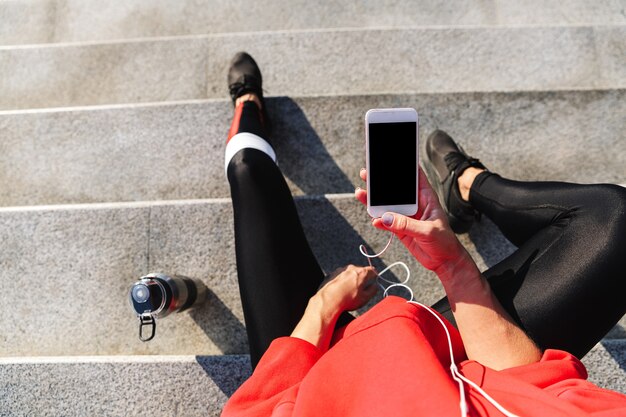 Top view of a young sportswoman holding mobile phone, listening to music with earphones, drinking water