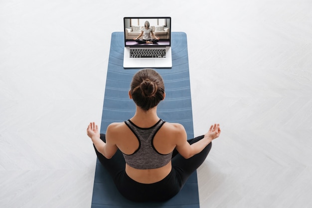 Vista dall'alto giovane donna sportiva utilizzando laptop per la formazione di hatha yoga con istruttore virtuale, facendo sukhasana