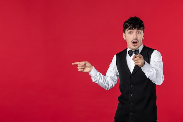 Top view of young surprised male waiter in a uniform with bow tie and pointing up something on the right side