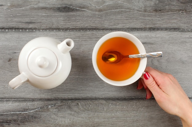 Top view, young woman hand with red nail holding cup of amber tea, with teapot next to it.