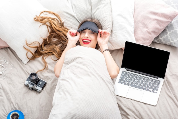 노트북, 전화, 사진 카메라를 들고 침대에 누워 있는 젊은 여성의 최고 전망. 퇴근 후 휴식