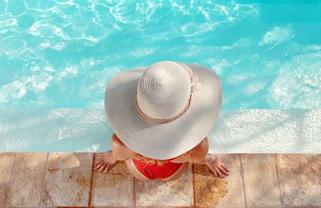 Top view of young woman in red one-piece swimsuit and straw hat relax near a swimming pool with legs in water