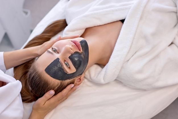 Top view on young woman uses the services of professional\
beautician at at spa. cropped unrecognizable master beautician puts\
black mask on client\'s face and gives a massage. health and\
longevity