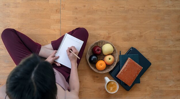 Photo top view of young woman writing on notebook with pencil while sitting on the floor at home.