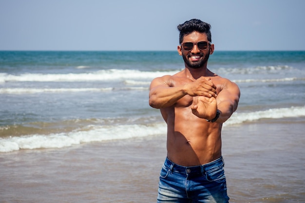 Topless atletico, muscoloso e sano uomo di colore warm up istruttore sulla spiaggia. modello di moda maschile indiano con pantaloncini di jeans jeans e corpo perfetto. cibo sano e vacanza attiva al mare