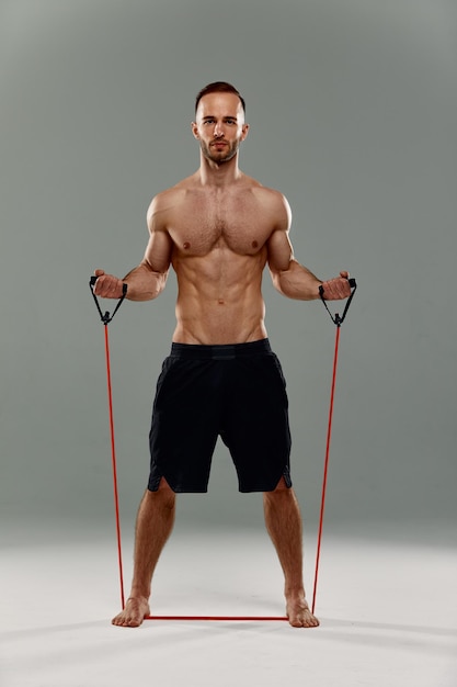 A topless sportsman performs fitness exercises with rubber band in a studio on gray background Bodybuilding Athlete