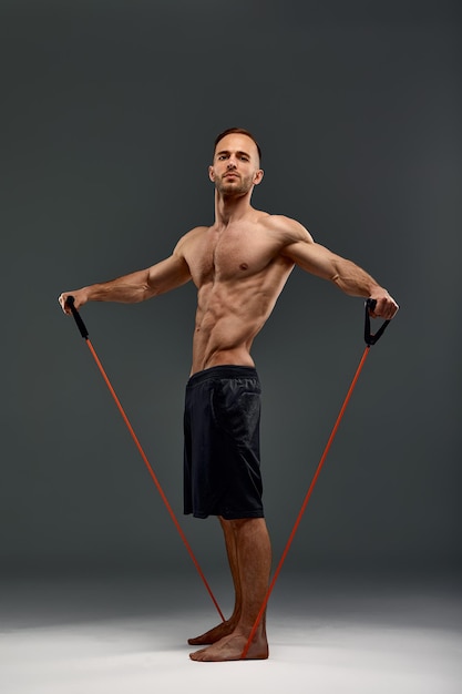 A topless sportsman performs fitness exercises with rubber band in a studio on gray background Bodybuilding Athlete