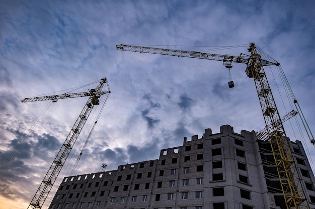 Torenkranen en onvoltooide hoge gebouwen met meerdere verdiepingen in aanbouw 's avonds met dramatische kleurrijke wolkenachtergrond