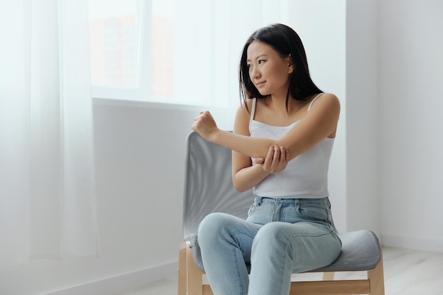 Tormented suffering tanned beautiful young asian woman hurting\
holding painful elbow at home interior living room injuries poor\
health illness concept cool offer banner