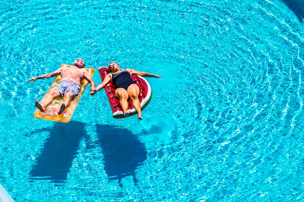 幸せな成熟したシニア退職者のための完全なリラックスは、夏の時間にプールの青い澄んだ水の上に着色されたリロスの膨脹可能なマットレスの上に横たわる