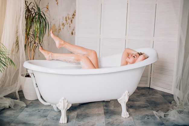 Total relaxation, purity, sensuality. Beautiful young caucasian with perfect soft skin showing her slender bare legs while lying in bathtub in modern spacious bathroom with green plants.