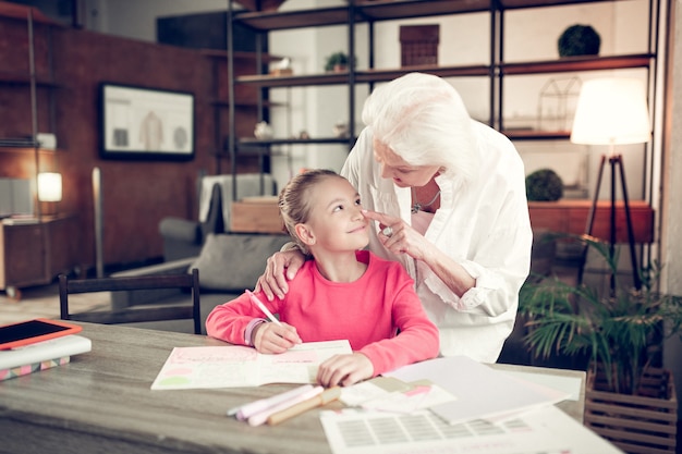 Touching nose. Grey-haired granny touching nose of her lovely cute girl doing homework at home