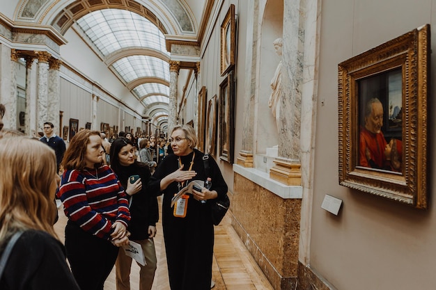 투어 가이드 여성은 루브르 박물관의 복도에서 작은 그룹의 젊은이들을 여행하며 이야기를 들려줍니다.