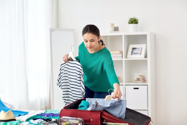 tourism, people and luggage concept - happy young woman packing travel bag at home or hotel room