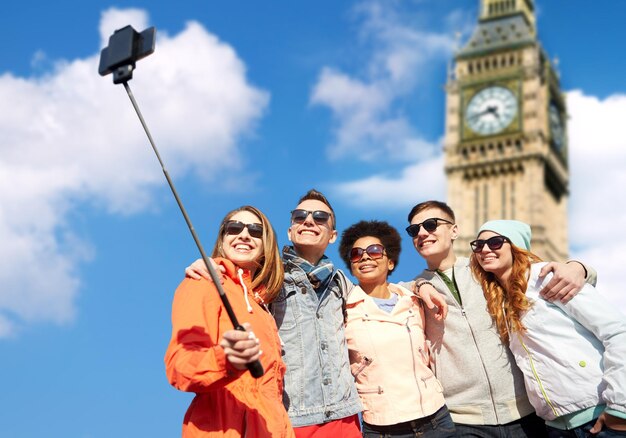 Tourism, Travel, People, Leisure And Technology Concept - Group Of Smiling Teenage Friends Taking Selfie With Smartphone And Monopod Over London Big Ben Tower Background