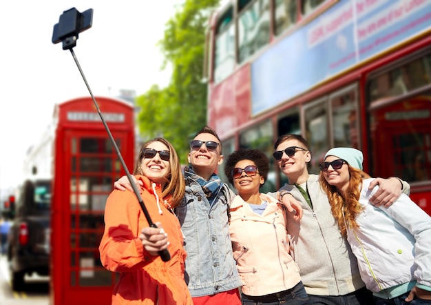 tourism, travel, people, leisure and technology concept - group of smiling teenage friends taking selfie with smartphone and monopod over london city street background