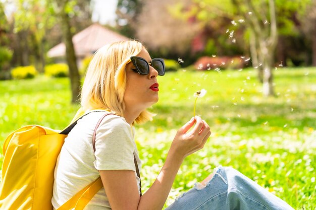 Una donna bionda turistica che soffia pianta di dente di leone che indossa un cappello e occhiali da sole seduto sull'erba in primavera accanto alle margherite in un parco della città